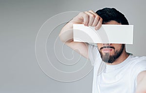 Young man isolated over background. Side view and selfie of bearded guy hide eyes from camera. Hold iece of paper or