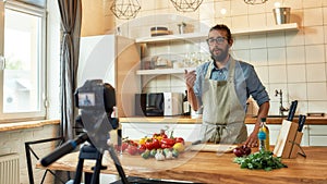 Young man, Italian cook in apron looking at camera, filming himself for culinary blog while preparing healthy meal with