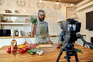 Young man, Italian cook in apron looking at camera, holding basil, filming himself for culinary blog while preparing