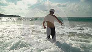 Young man in jeans enters shallow sea water with his back to camera in slow motion. The waves roll on shore. Summer at