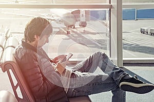 Young man in jeans and jacket sits on chair spend time by using mobile phone in airport lounge. Booking hotel in foreign country.