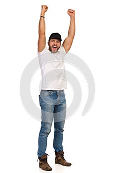 Young Man In Jeans, White T-shirt And Black Cap Is Standing With Arms Raised And Shouting