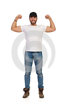 Young Man In Jeans, White T-shirt And Black Cap Is Standing And Flexing Muscles