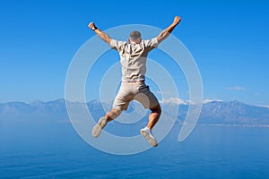 Young man jumping on rock with his hands up blue ocean mountain peaks horizon. Active holiday adventure, tourism action, healthy