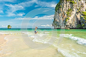 Young man jumps in the shallow sea waters with a large limestone cliff covered with tropical vegetation