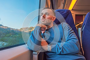 Young man keeps his hand on his chin and looks out the train window from the passenger compartment, enjoying the view of nature