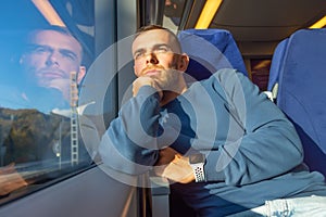 Young man keeps his hand on his chin and looks out the train window from the passenger compartment, enjoying the view of nature