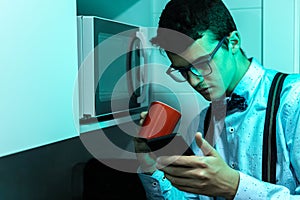 Young man in a kitchen drinking a red cup of coffee with milk and looking at the messages on the mobile phone while having