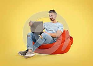 Young man with laptop on bean bag chair, yellow background