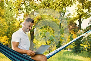 Young man with laptop in comfortable hammock at garden