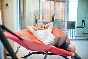 Young man with laptop holding hands behind head, relaxing and dreaming while lying on a hammock in office space. Taking time for a