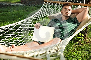 Young man with laptop resting in comfortable hammock at garden