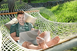 Young man with laptop resting in comfortable hammock at garden