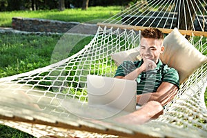 Young man with laptop resting in comfortable hammock at garden