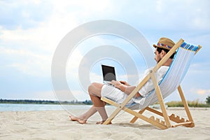 Young man with laptop sitting in deck chair on sea beach