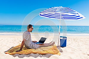 Young man with laptop working on the sand beach under umbrella on sea cost