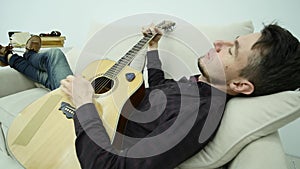 The young man lays on the couch and plays the guitar