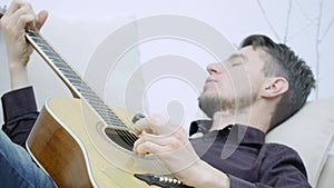 The young man lays on the couch and plays the guitar