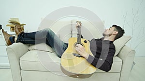 The young man lays on the couch and plays the guitar