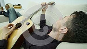 The young man lays on the couch and plays the guitar