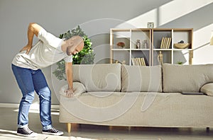 Young man leaning on sofa at home is holding his lower back due to severe pain and cramps.