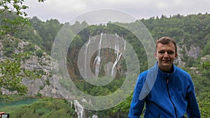 A young man leans on a wooden railing. In the background waterfalls in Plitvice Lakes National Park. Teenager in a blue hoodie