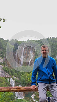 A young man leans on a wooden railing. In the background waterfalls in Plitvice Lakes National Park. Teenager in a blue hoodie