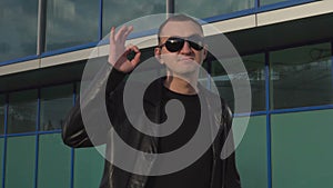 Young man in leather jacket and sunglasses standing outdoor and showing okay