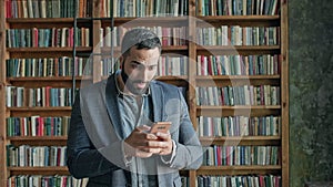 Young Man In The Library With Mobile Phone In His Hands. He Is Emotionally Playing Game On The Phone.