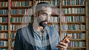 Young Man In The Library With Mobile Phone In His Hands. He Looks And Closes One Eye. Stylish Guy With Beard In Jacket.