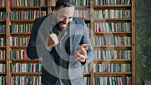 Young Man In The Library With Mobile Phone In His Hands. Something Pleasantly Surprises Him.