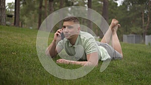 A young man lies on the grass and talks on the phone. A student in the park rests on the grass with communicates by