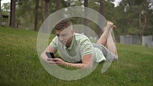 A young man lies on the grass and texts in a smartphone. Satisfied man is resting in nature with a phone.