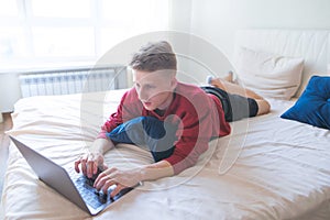 Young man lies at home on the bed and uses a laptop. Freelancer works on a laptop at home in a cozy room