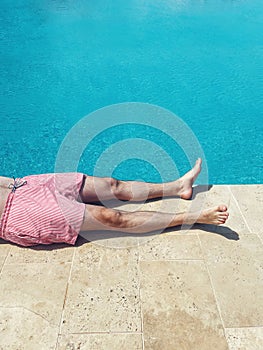 A young man lies on poolside at resort hotel during vacation. Close up of male legs near turquoise water