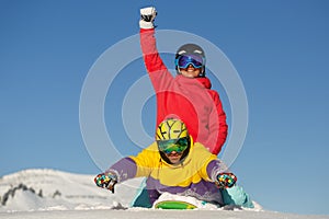 A young man lies on a snowboard in a superman pose, a girl sits on a guy with a raised hand