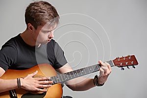 Young man with light beard playing acoustic guitar, on grey