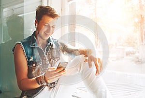 Young man listen music with headphones and smartphone sitting on windowsill