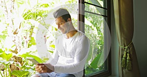 Young Man Listen To Music On Tablet Computer With Earphones Sitting One On Window Sill Looking To Tropical Garden