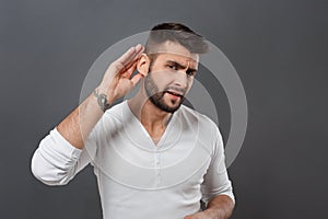 Young man listening holding hand near ear over grey background.