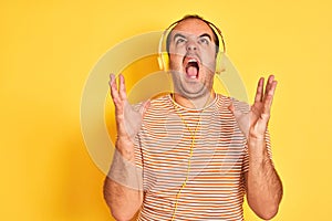 Young man listening to music using headphones standing over isolated yellow background crazy and mad shouting and yelling with