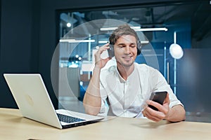 A young man listens to podcasts in headphones, holds a mobile phone in his hands, talks on video communication. Sitting at work in