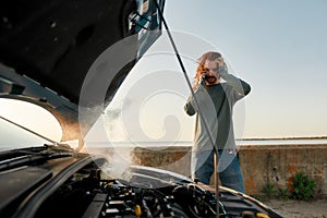 Young man looking desperate, using smartphone, calling for assistance while standing near his broken car with open hood