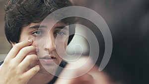 Young man is looking at face and touching skin under eyes with finger in front of mirror