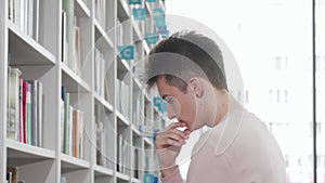 Young man looking thoughtfully at the bookshelves at the library