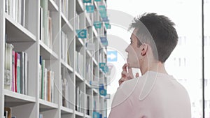 Young man looking thoughtfully at the bookshelves at the library