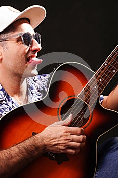 Young man looking to the side sticking out his tongue, playing guitar with black sunglasses, white cap