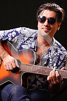 Young man looking towards the camera smiling, playing guitar, with black sunglasses and blue and white hawaiian shirt