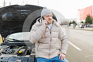 A young man looks angry and frustrated near his broken car and speaks on his mobile phone,