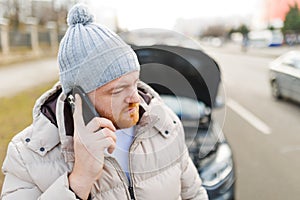 A young man looks angry and frustrated near his broken car and speaks on his mobile phone,